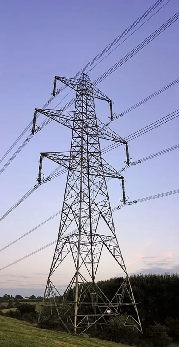 High-voltage steel transmission tower standing in a rural landscape at sunset, with multiple power lines extending across the sky.